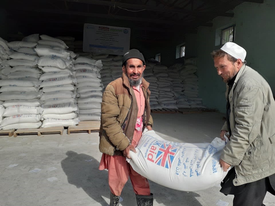 A man called Tora is pictured wearing a a peach-coloured tunic with matching trousers, a brown puffer jacket, black beanie hat and black wellington boots. He is smiling at the camera holding one end of a white sack of wheat seed whilst another man holds the other end. In the background there are dozens of the white sacks stacked on top of each other.